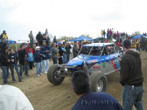 1990 Ford Bronco - blue single-seat off-road race buggy navigates a dirt course closely lined by a large crowd of spectators.