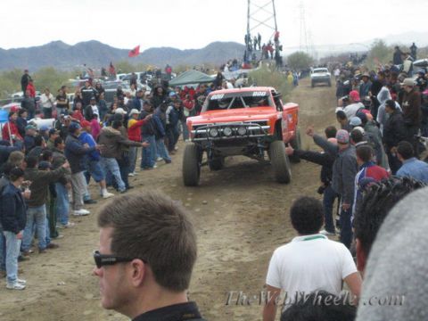 1990 Ford Bronco - red off-road race truck marked 800 drives a dirt course flanked by a large cheering crowd with desert mountains behind.