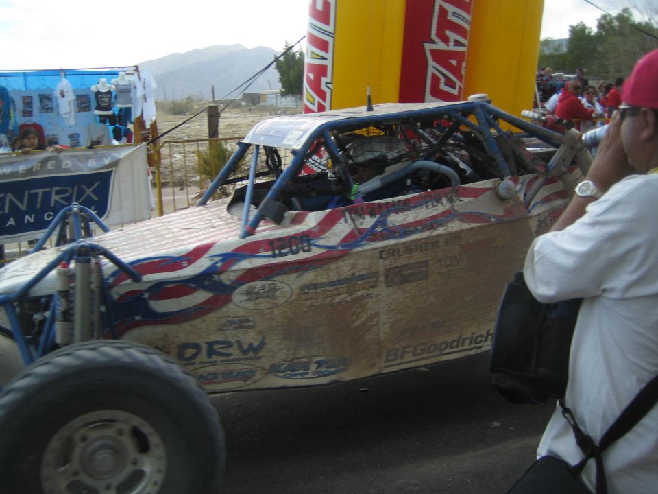 Tube-frame off-road race buggy with US-flag graphics passing spectators at a desert finish