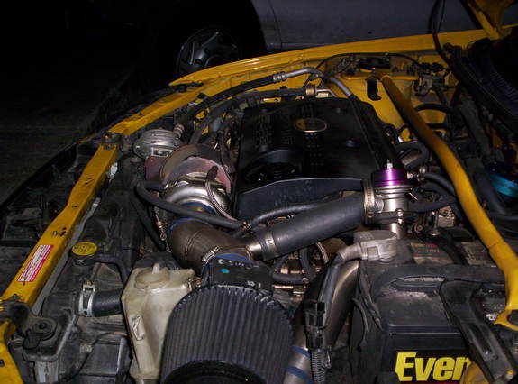 Turbocharged engine bay of a yellow car with cone air filter, purple blow-off valve, and Everstart battery