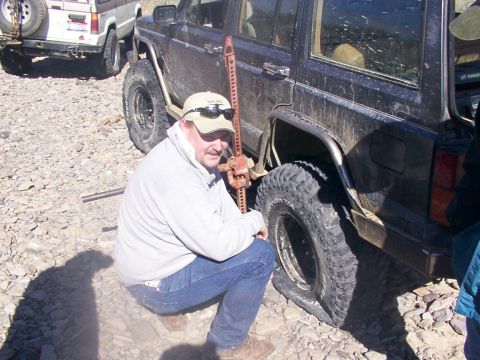 1978 Ford Bronco - A man uses a hi-lift jack to change a flat tire on a dark off-road SUV, with another vehicle visible in the background.