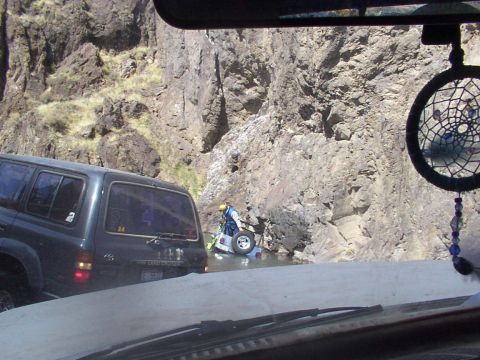 1978 Ford Bronco - A dark SUV navigates a rocky terrain beside a river where a person is attending to a vehicle partially submerged in the water.