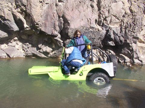 1978 Ford Bronco - Two people are in a bright green Jeep that is partially submerged in a body of water, with a rocky cliff behind them.