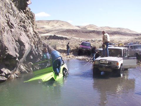 1978 Ford Bronco - Several off-road vehicles are partially submerged in a shallow stream, with people interacting around them in a , arid landscape.