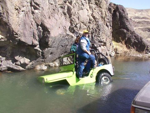 1978 Ford Bronco - A green Jeep CJ-5 navigates through shallow water near a rocky cliff face.
