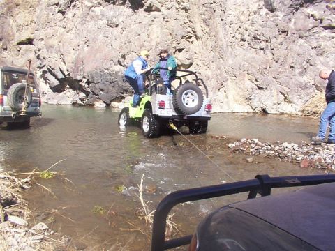 1978 Ford Bronco - Several off-road vehicles navigate a shallow riverbed with rocky banks under a clear sky.