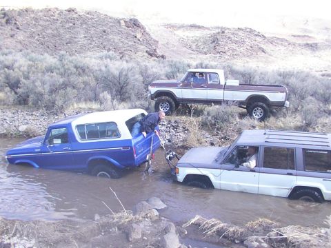 1978 Ford Bronco - Three off-road vehicles are stuck in a shallow stream, with one person attempting to assist.