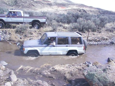1978 Ford Bronco - A white and grey SUV navigates through a shallow stream, with a pickup truck assisting from the bank.