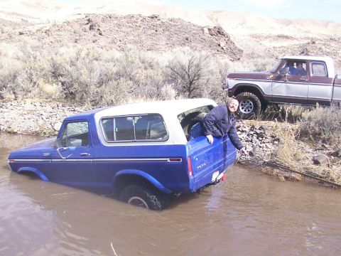 1978 Ford Bronco - A blue Ford Bronco is stuck in shallow water while a person watches from the rear, with another vehicle nearby.