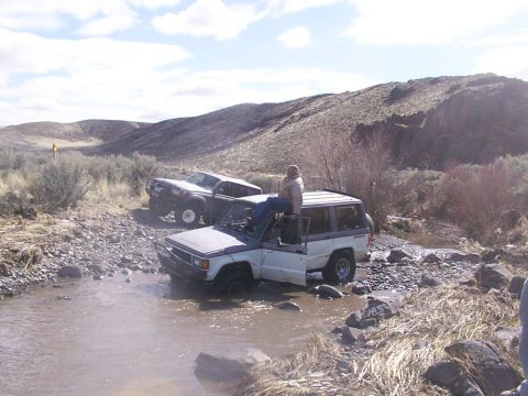 1978 Ford Bronco - Two SUVs navigate a shallow, rocky stream in a dry, hilly landscape under a cloudy sky.