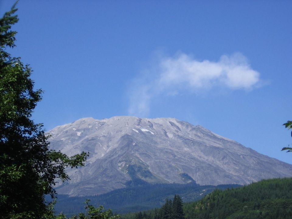 Gray volcanic mountain with steam or cloud at summit under blue sky, framed by evergreen trees
