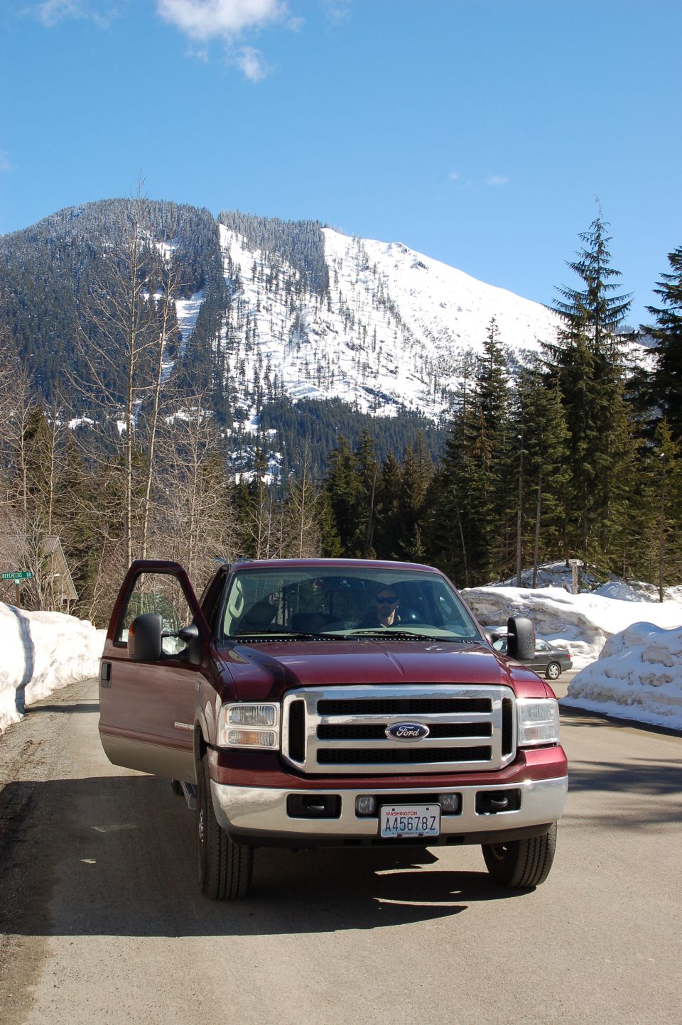Maroon Ford F-250 truck with driver's door open, parked on a dirt road with snow on the sides. Mountains in background