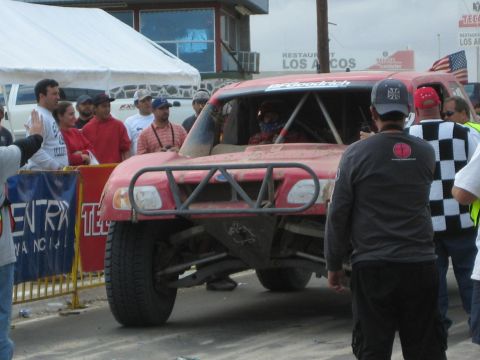 Front view of a dusty red tube-frame off-road race truck stopped near a Los Arcos restaurant