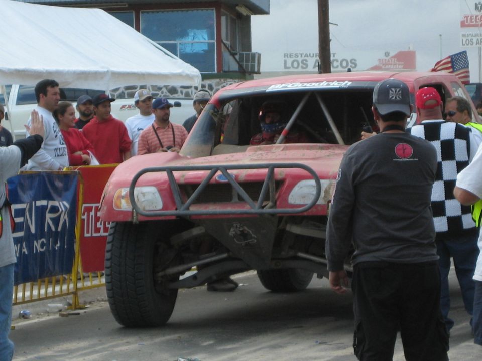 Front view of a dusty red tube-frame off-road race truck stopped near a Los Arcos restaurant