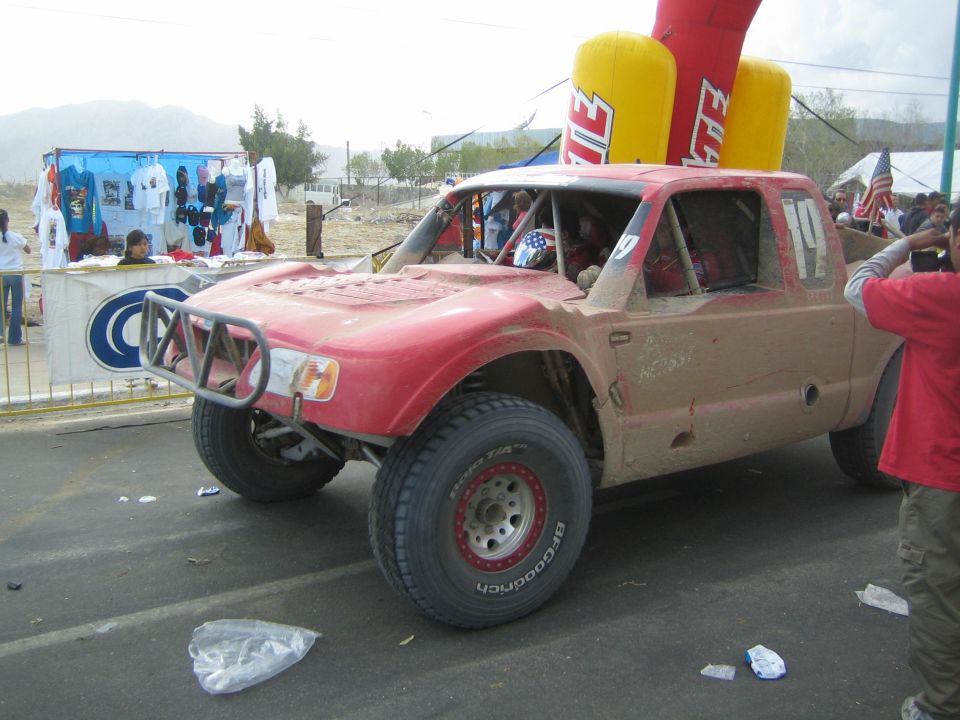Number 110 dusty red off-road race truck under an inflatable arch with spectators