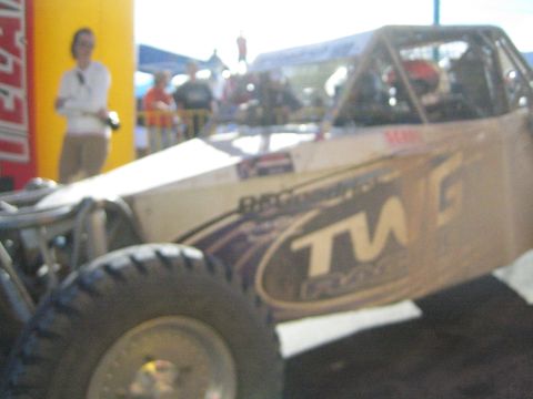 Blurry tan tube-frame off-road buggy with TW graphics near a vendor tent