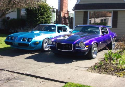 1979 Pontiac Trans Am - Two vintage muscle cars, one blue and one purple, are parked side-by-side in a residential driveway.