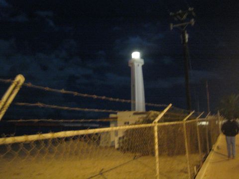 Illuminated white lighthouse behind a chain-link and barbed-wire fence at night