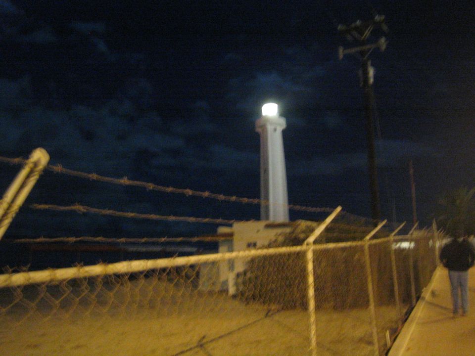 Illuminated white lighthouse behind a chain-link and barbed-wire fence at night