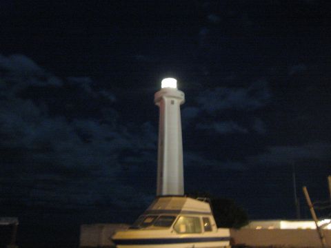 Tall lit white lighthouse rising above a stored cabin boat at night