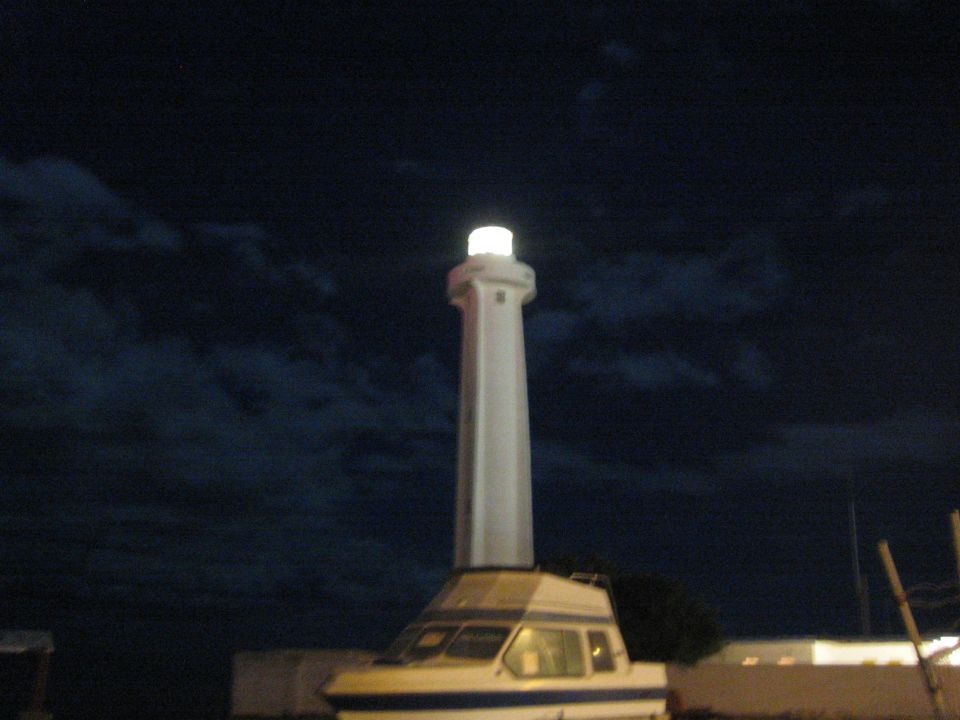 Tall lit white lighthouse rising above a stored cabin boat at night