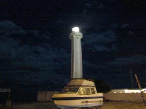 Illuminated lighthouse behind a small cabin boat on dry ground at night
