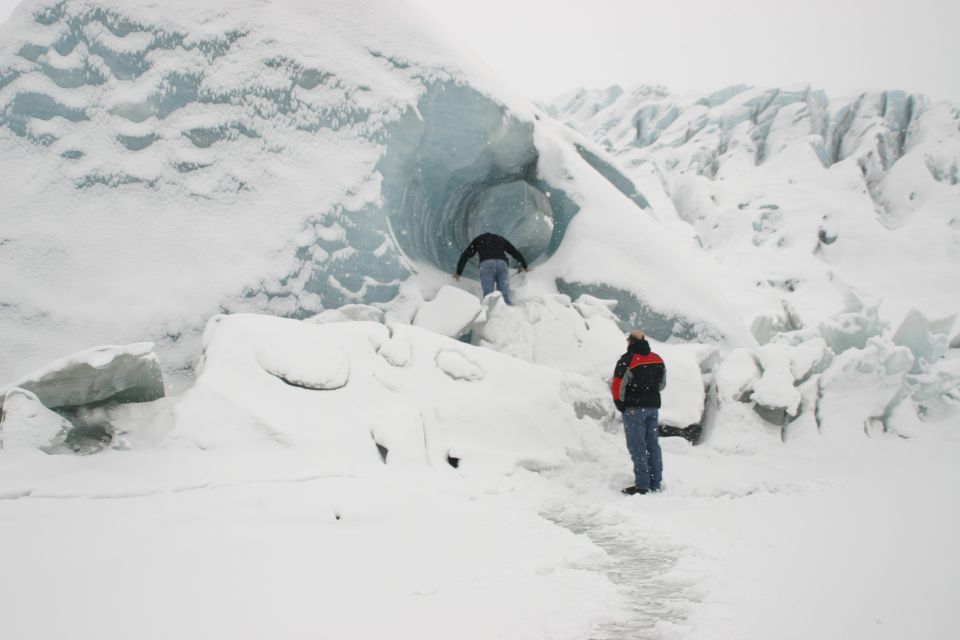 Two people stand near a snow-covered glacier with a large ice cave entrance. One person enters the cave