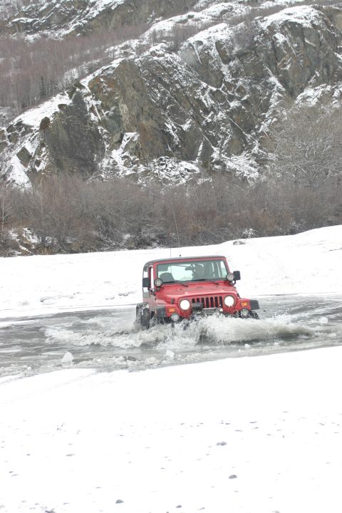 2003 Jeep TJ - A red 2003 Jeep TJ navigates through icy water, with snow-capped mountains providing a dramatic backdrop.