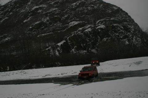 2003 Jeep TJ - Two red SUVs navigate a snowy road near a mountain, kicking up slush.