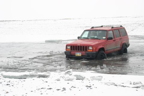 2003 Jeep TJ - A red Jeep Cherokee navigates through shallow, icy water with snow-covered ground.