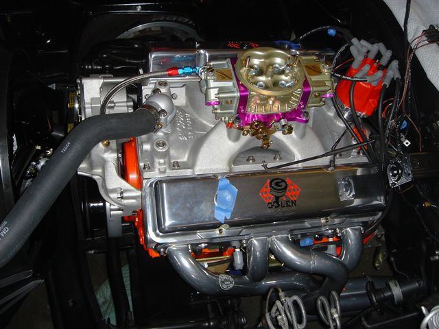 Close-up of a V8 engine with a Holley carburetor, chrome valve covers, and custom headers. There's a 'QOLEN' logo on one of the valve covers
