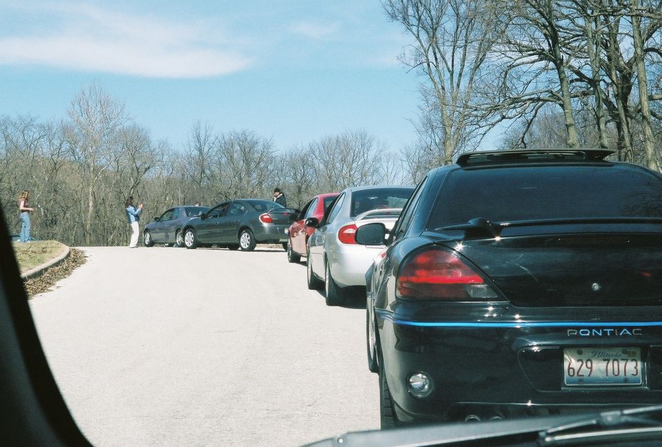 Several cars parked on a road with trees in the background. A black Pontiac is in the foreground