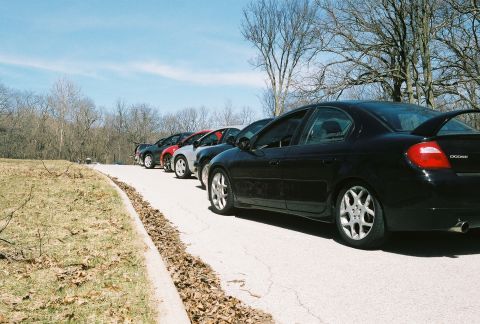 2004 Dodge SRT-4 - A line of cars, including a black Dodge Neon SRT-4, are parked on a paved road with trees in the background.