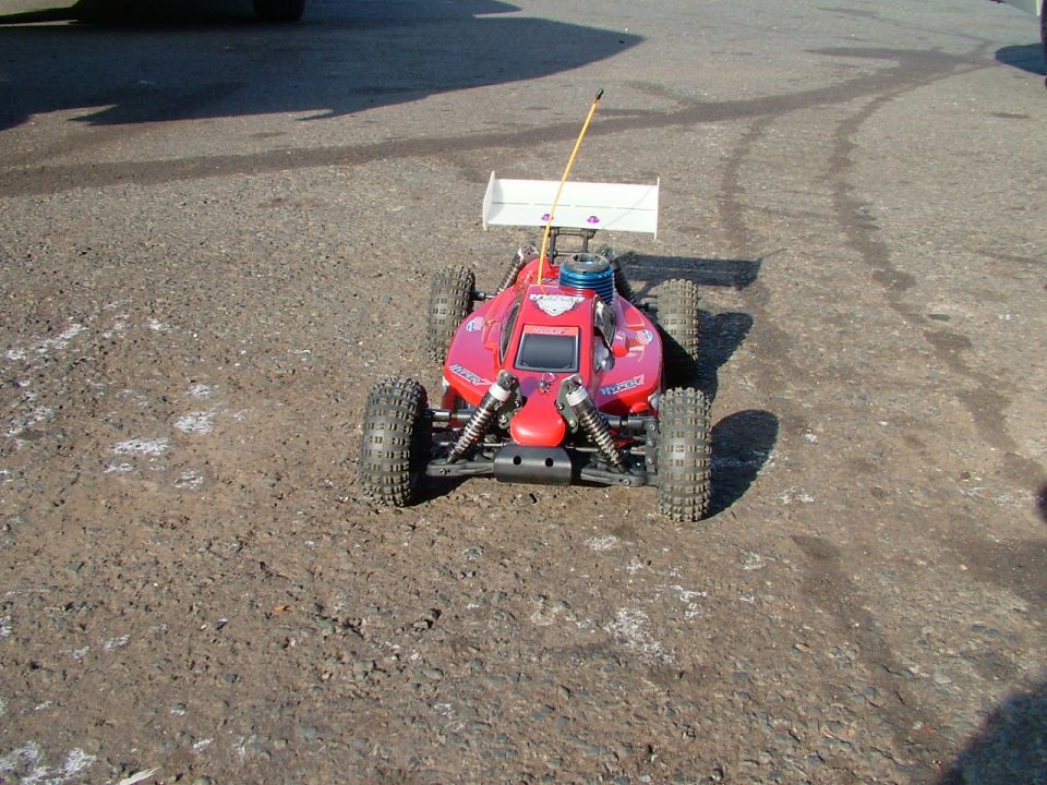 Red remote control buggy with a white spoiler and an antenna, sitting on a dirt and asphalt surface