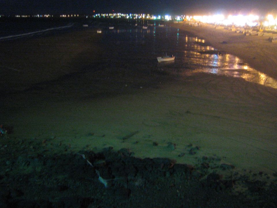 Wet sandy beach at night with a single boat and distant city lights