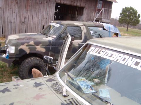1988 Ford Bronco - Two camouflaged vehicles are parked in front of a wooden barn, one with its door ajar.