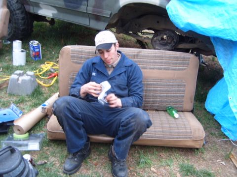 1988 Ford Bronco - A person sits on a vehicle seat amidst various automotive parts and supplies, with a tarp covering part of the scene.