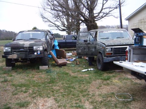 1988 Ford Bronco - Two Ford Broncos are parked in a yard, one with its door open, surrounded by parts and a pickup truck.