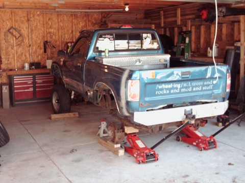 1977 Ford F-150 - A blue pickup truck is elevated on jacks inside a garage, undergoing maintenance or modification.