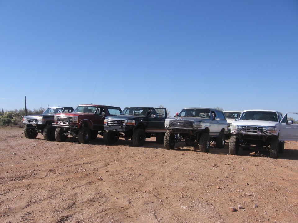 Several lifted Ford Broncos and F-Series trucks parked in a dirt lot under a clear blue sky