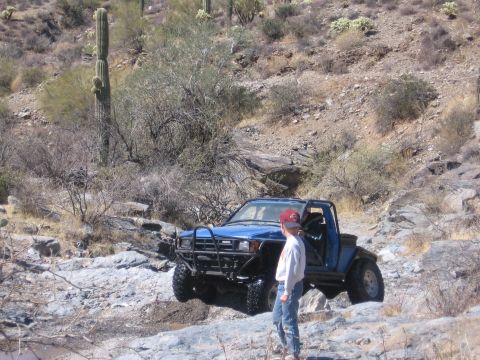1990 Ford Bronco - A blue pickup truck modified for off-road use navigates a rocky trail in a desert environment.