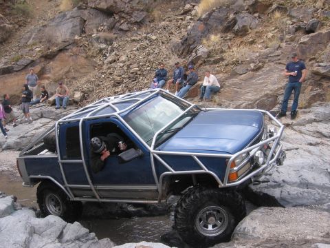 1990 Ford Bronco - A heavily modified blue truck navigates a rocky stream bed with onlookers observing the off-road challenge.