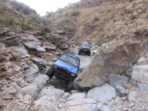 1990 Ford Bronco - Multiple off-road vehicles traverse a challenging, rocky mountain pass under a bright sky.
