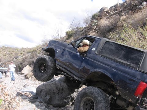 1990 Ford Bronco - A blue 1990 Ford Bronco navigates a rocky off-road course with its driver visible.