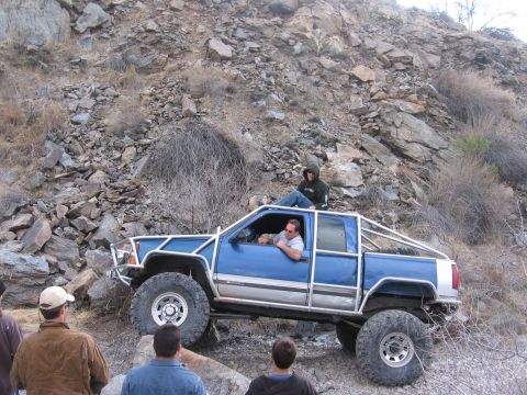 1990 Ford Bronco - Modified blue pickup truck navigating a rocky trail with passengers inside and on top.