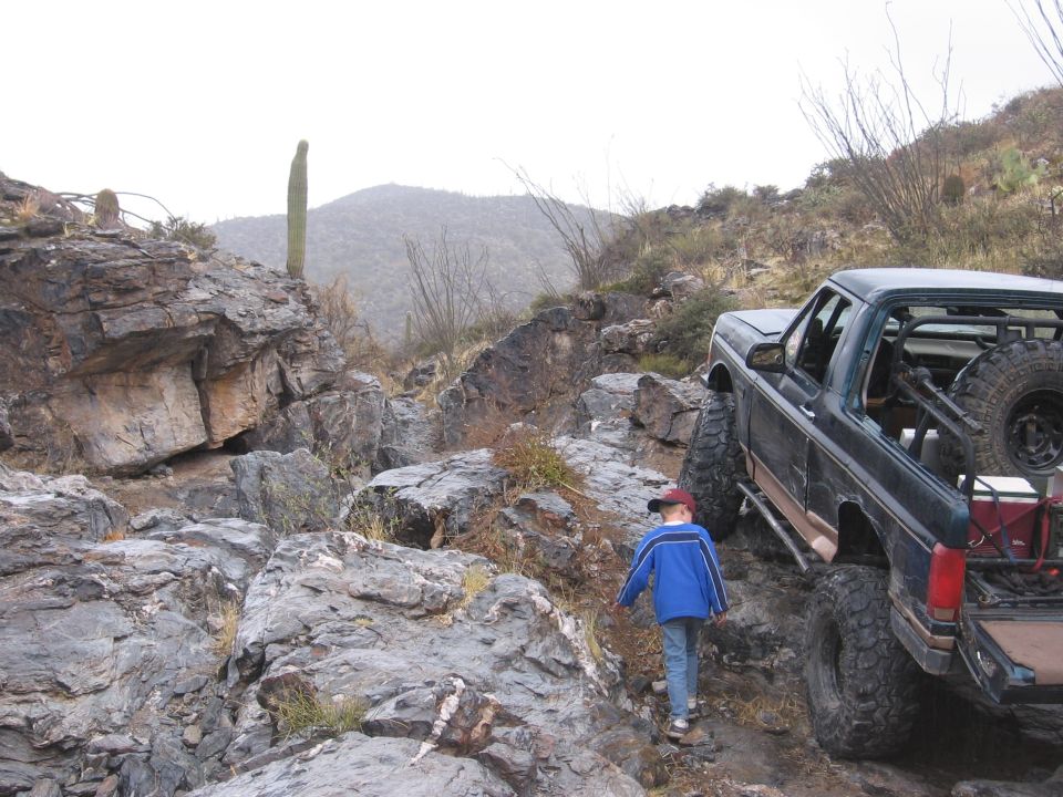 Dark green Ford Bronco with lifted suspension and spare tire on the back, driving on a rocky desert trail