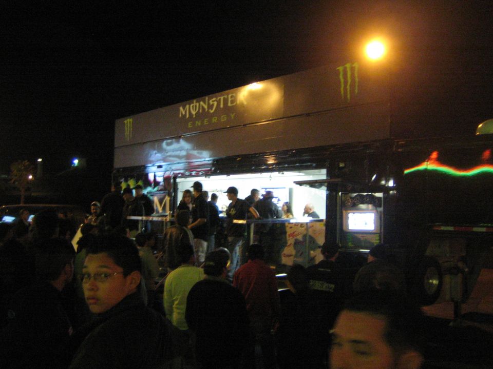 Crowd around a Monster Energy trailer booth under a street lamp at night