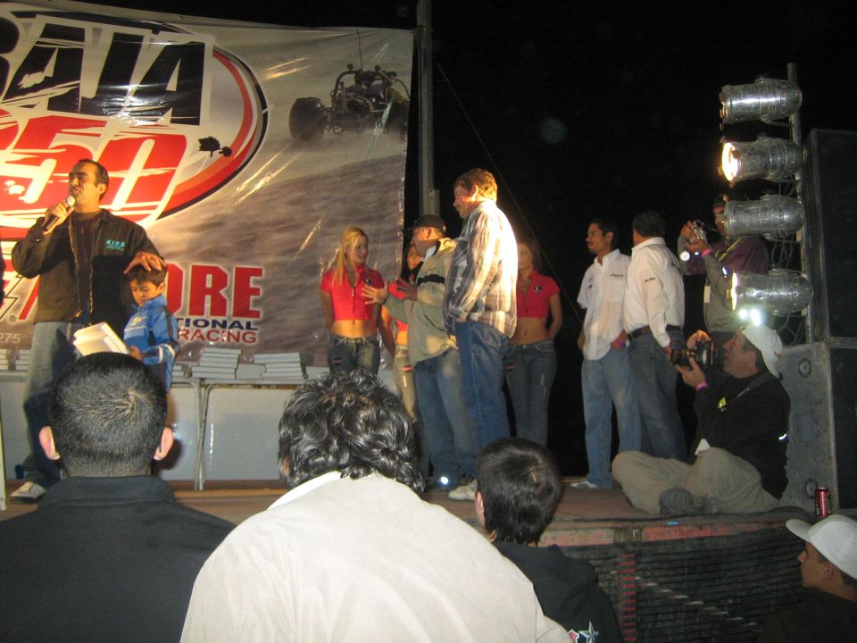 Host with mic on a Baja 250 stage at night addressing a crowd
