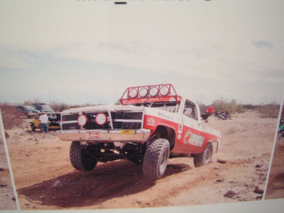 Photo of a red and white off-road race truck with roof light bar climbing a desert berm
