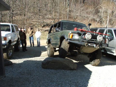 1996 Ford Bronco - An off-road SUV is positioned with one tire on a large rock, demonstrating its articulation. Another SUV and several people are visible in…
